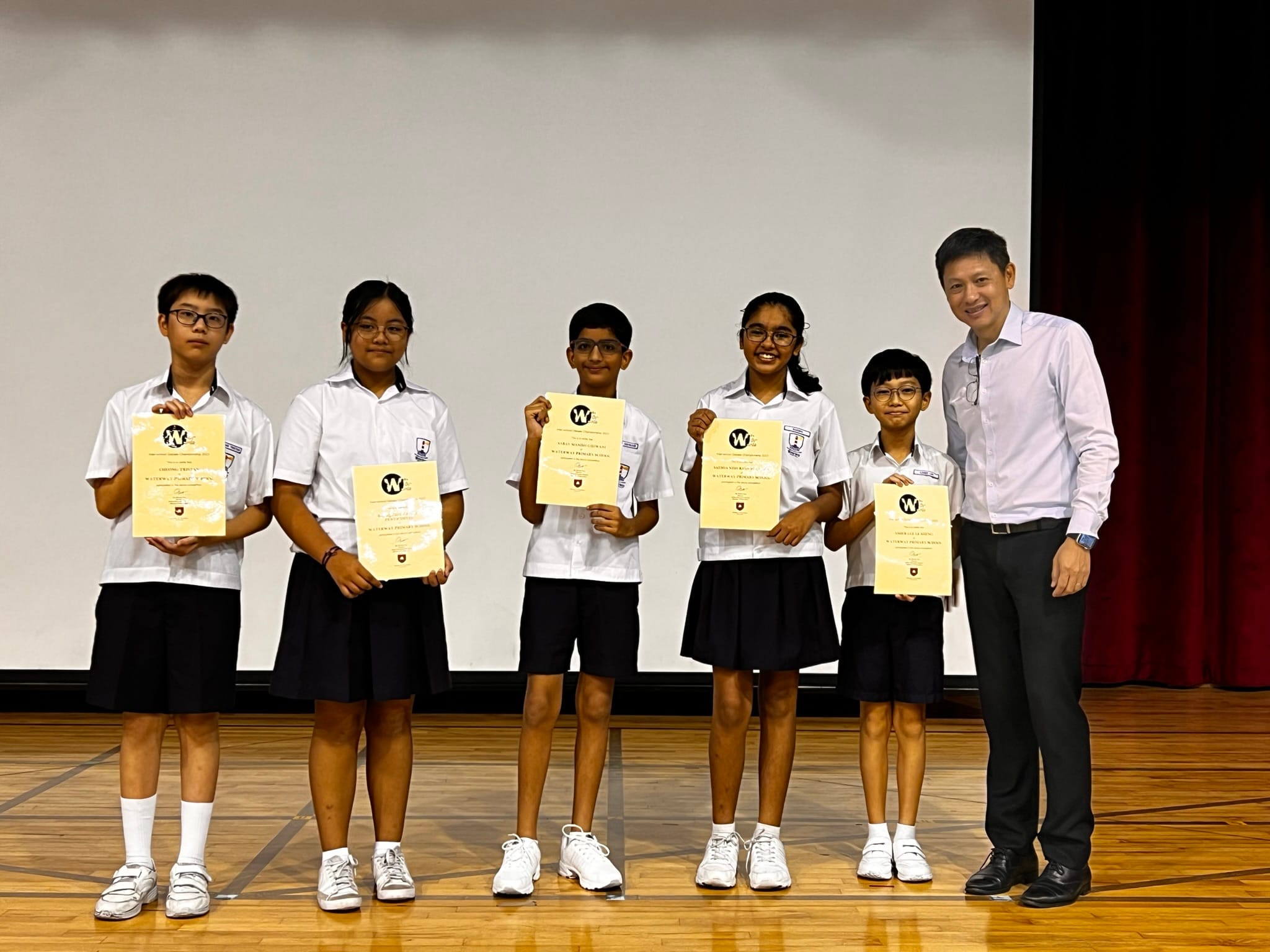 Five students in white uniforms hold certificates on a stage, standing beside the Principal in a light shirt.